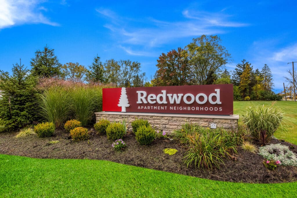 A landscaped area with a stone sign that reads "Redwood Apartment Neighborhoods" in front of grass, trees, and a blue sky.