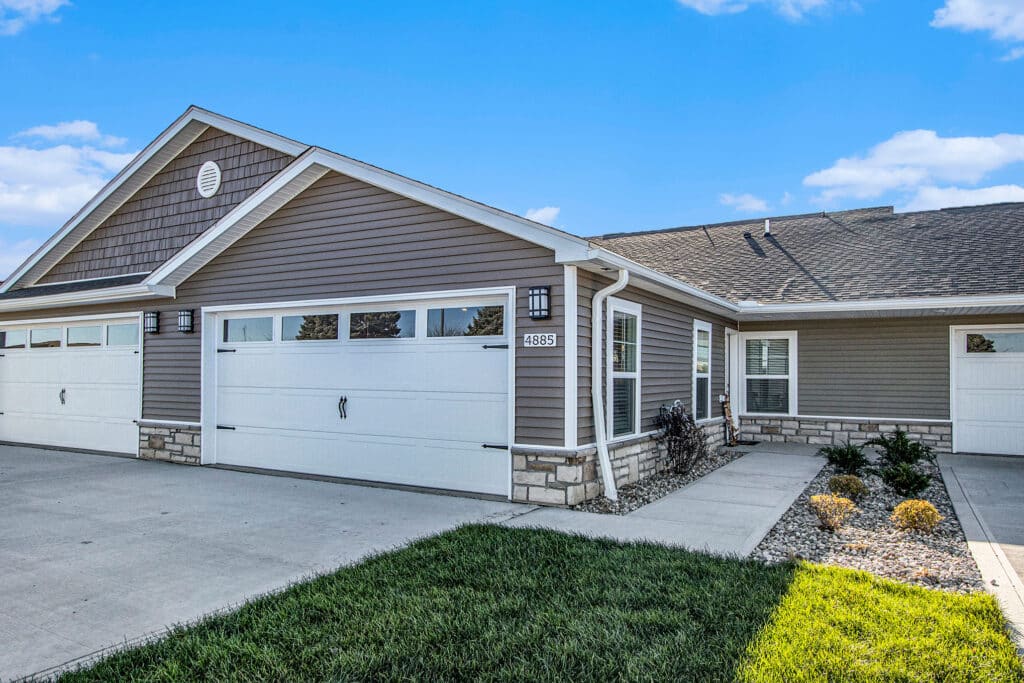 Single-story suburban house with beige siding, white trim, a double garage, paved driveway, walkway, and minimal landscaping under a blue sky.