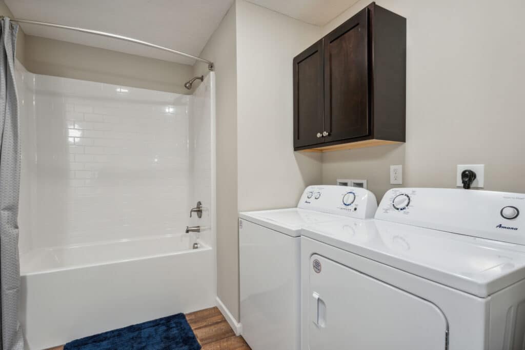 Laundry area with a washing machine and dryer next to a bathroom with a white bathtub, shower curtain, dark cabinet, and blue rug on wood flooring.