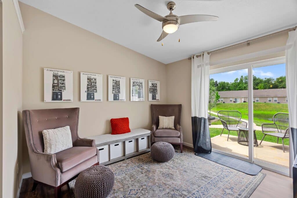 A bright sitting room with two armchairs, a storage bench, poufs, and framed wall art, featuring large windows and a view of a patio with outdoor seating and greenery.