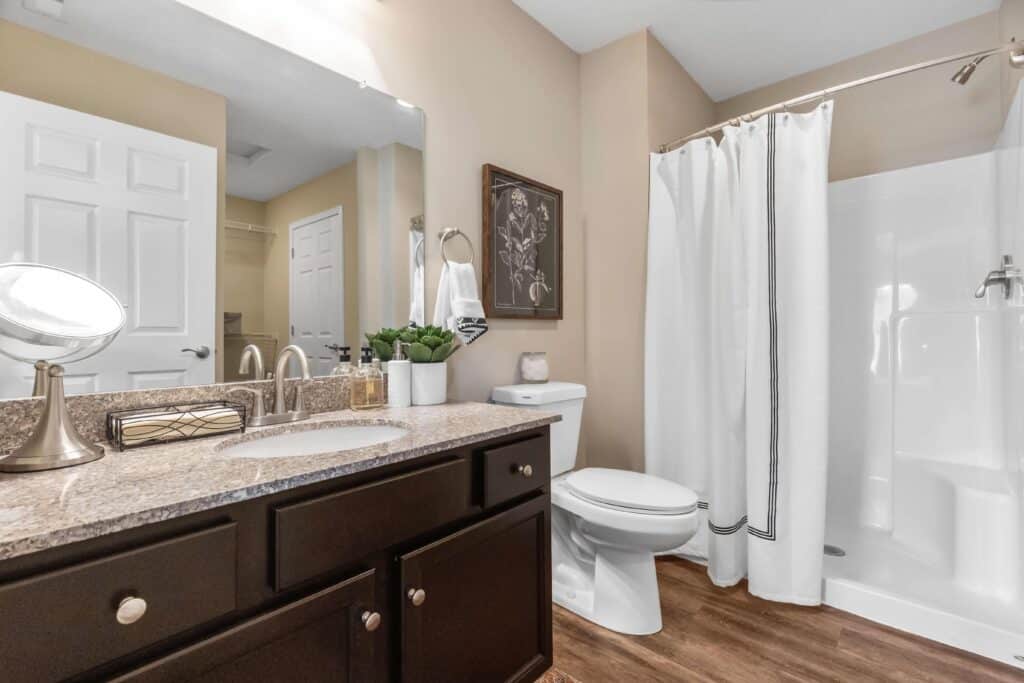 Modern bathroom with a granite countertop, dark wood cabinets, white toilet, walk-in shower with white curtain, and beige walls. A framed picture and plants decorate the space.
