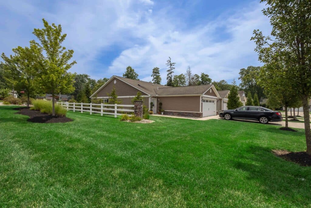 Single-story house with a two-car garage, white fence, black car parked in driveway, and well-maintained green lawn under a partly cloudy sky.