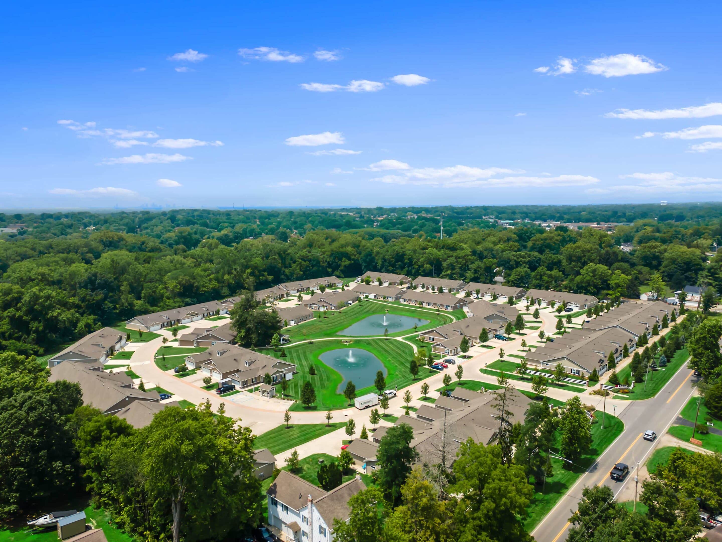 Aerial view of a suburban neighborhood with houses arranged in a circular pattern around a central green park with two fountains. Trees and roads surround the area under a blue sky.