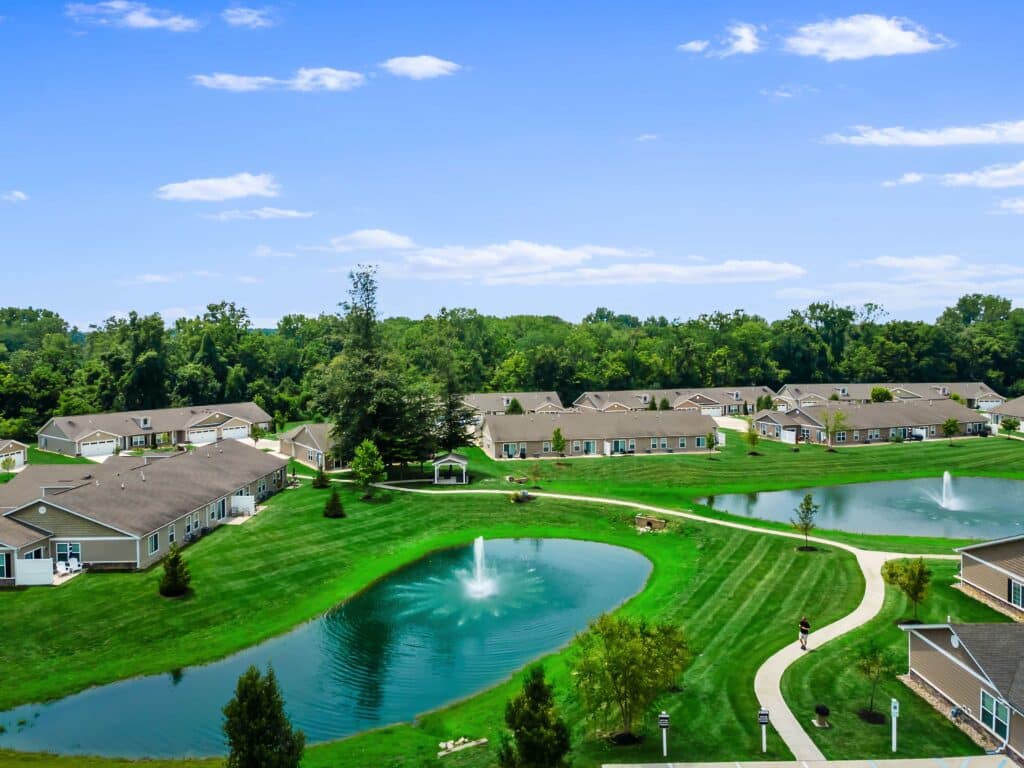 A residential community with single-story homes, manicured lawns, winding paths, small ponds, and fountains, surrounded by trees under a blue sky.