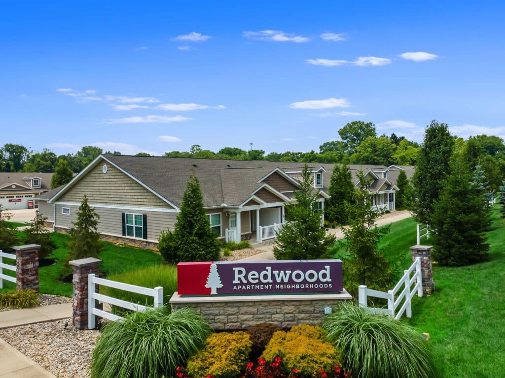 Single-story apartment buildings with attached garages and landscaped grounds, featuring a sign that reads "Redwood Apartment Neighborhoods" in the foreground.