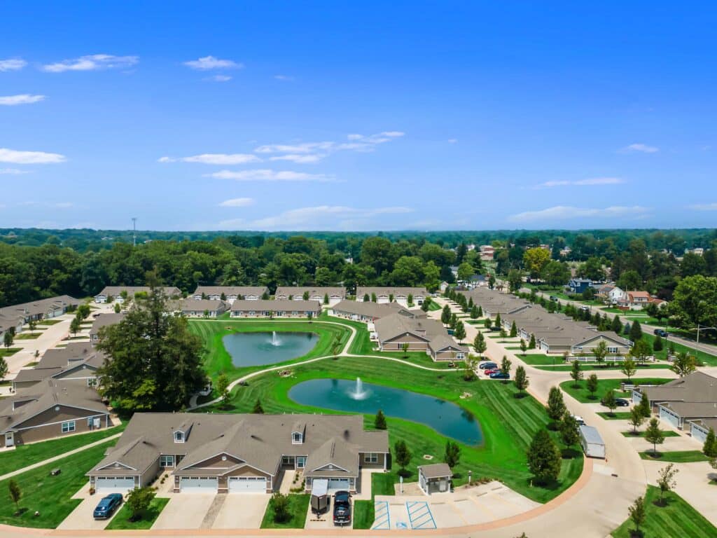 Aerial view of a suburban neighborhood with single-story houses, ponds, green lawns, trees, and driveways under a clear blue sky.