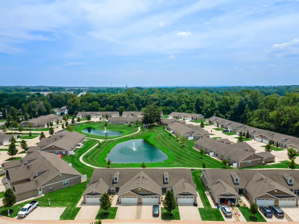Aerial view of a suburban neighborhood with rows of houses, two small ponds with fountains, and green lawns surrounded by trees under a blue sky.