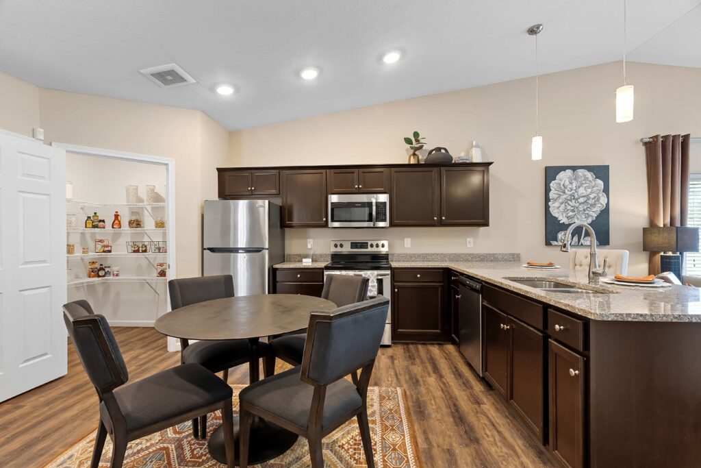 Modern kitchen with dark wood cabinets, stainless steel appliances, a round dining table with four chairs, granite countertops, and an adjacent walk-in pantry.