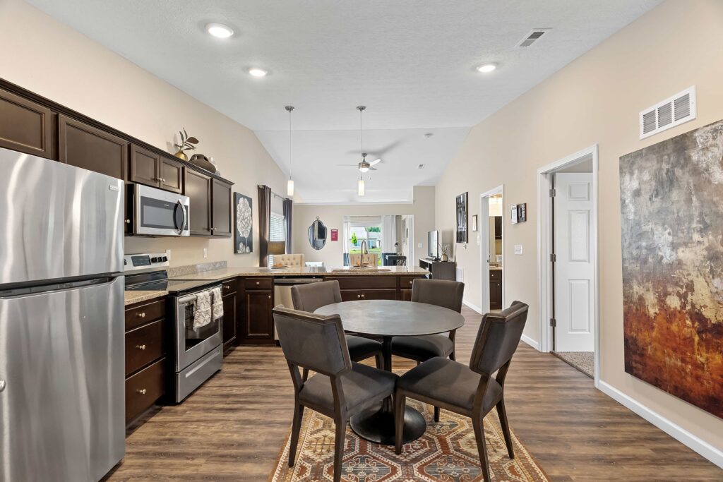 Modern kitchen and dining area with dark cabinets, stainless steel appliances, round table with four chairs, hardwood flooring, and wall art. Living area visible in the background.