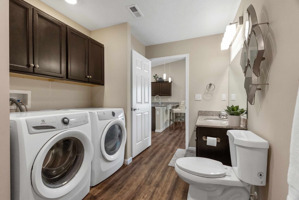 Laundry room with washer and dryer, dark cabinets, toilet, sink with mirror, and open door showing adjacent room with desk and chair.