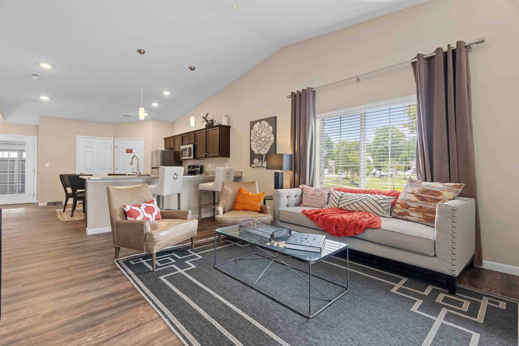 Modern living room with a gray sofa, armchair, coffee table, and kitchen in the background; large window with curtains lets in natural light.