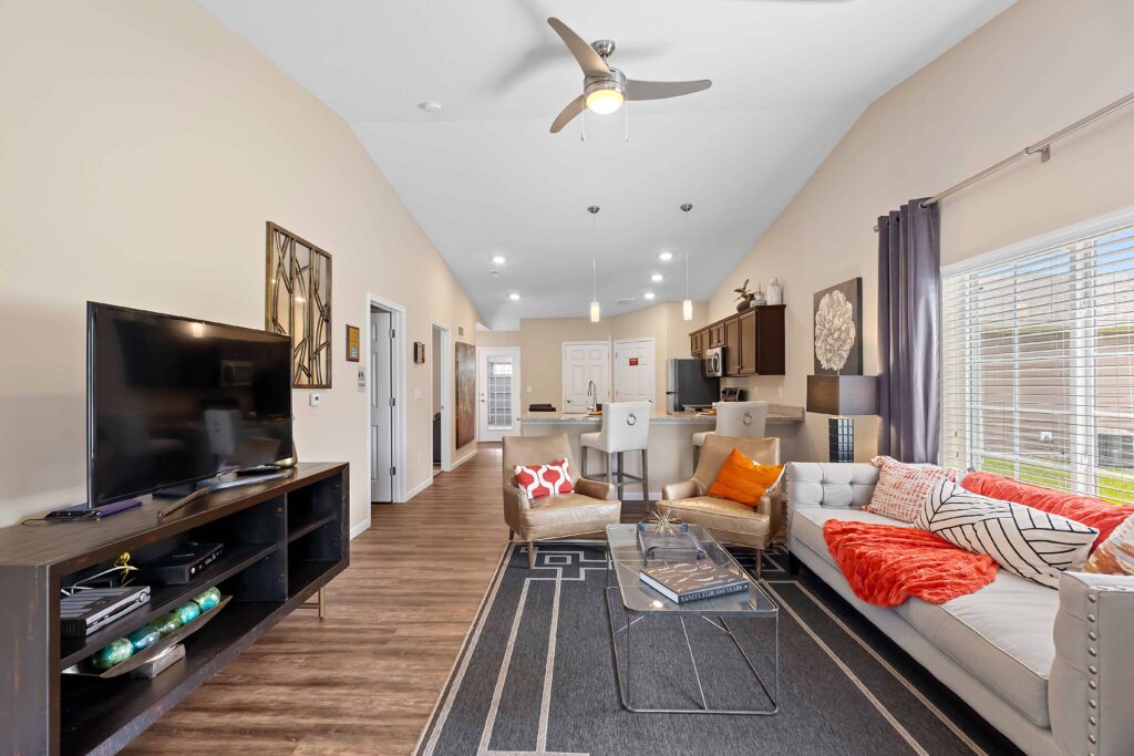 Modern living room with a TV, sofa, colorful pillows, glass coffee table, dining area, and kitchen in an open-concept space with wood flooring and large windows.