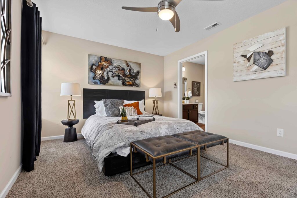 A modern bedroom with a neatly made bed, two lamps, abstract wall art, a bench at the foot of the bed, and an open doorway leading to a bathroom.