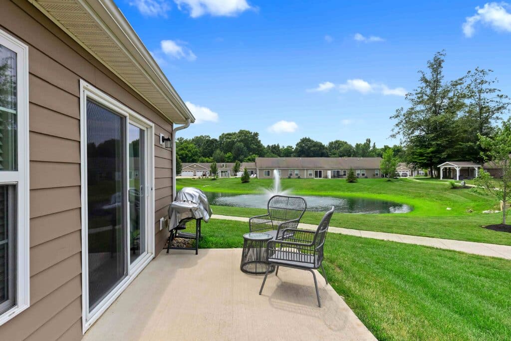 Patio with metal chairs and a grill overlooks a grassy yard and pond, with residential buildings and trees in the background under a blue sky.