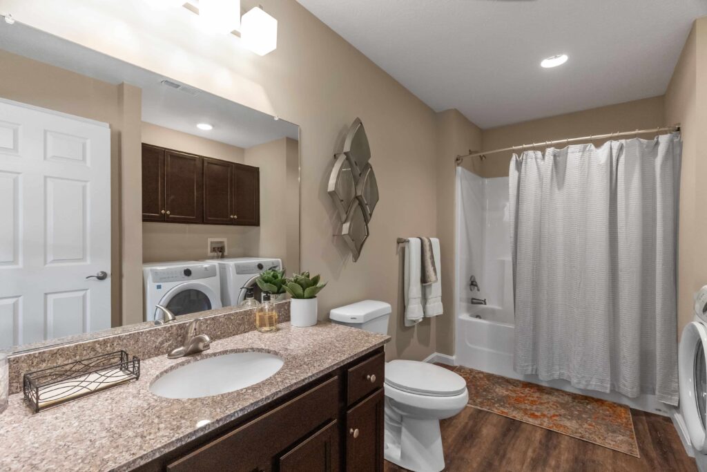 A bathroom with a sink, toilet, bathtub with shower curtain, washer and dryer, brown cabinets, and decorative plants on the countertop.