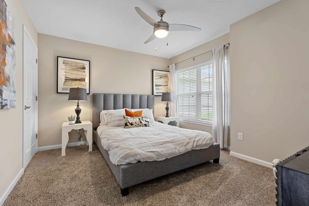 A neatly made bed with a gray upholstered headboard is centered between two white nightstands in a carpeted bedroom with beige walls and a ceiling fan.