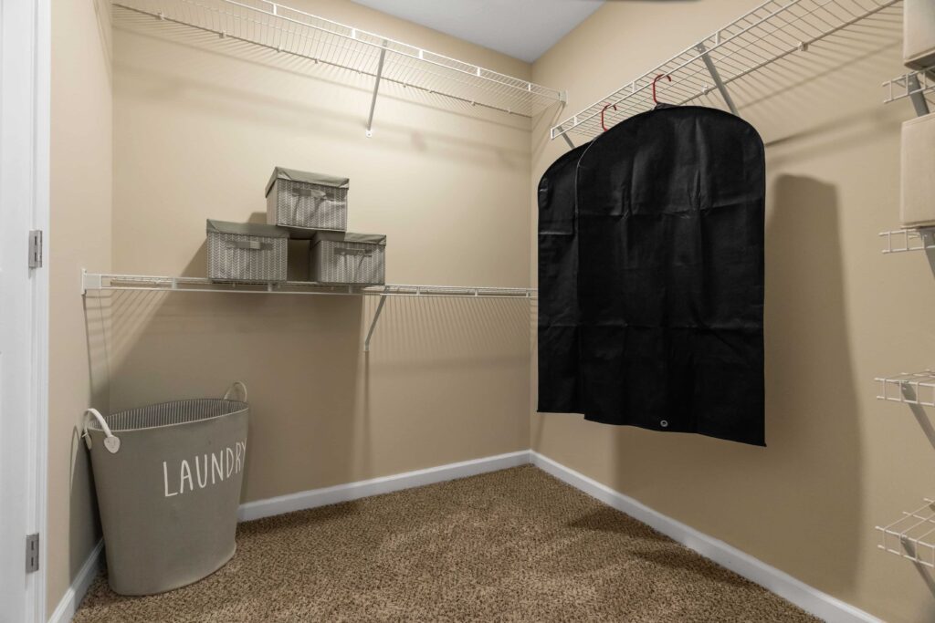 A walk-in closet with beige walls, wire shelves, storage bins, a black garment bag, and a large laundry basket on carpeted floor.