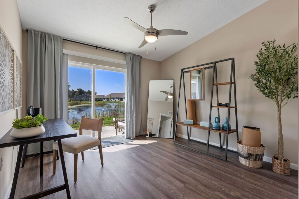 Bright home office with a desk, chair, plant, large shelving unit, and floor mirror. Sliding glass doors open to a patio with a view of a pond and houses.