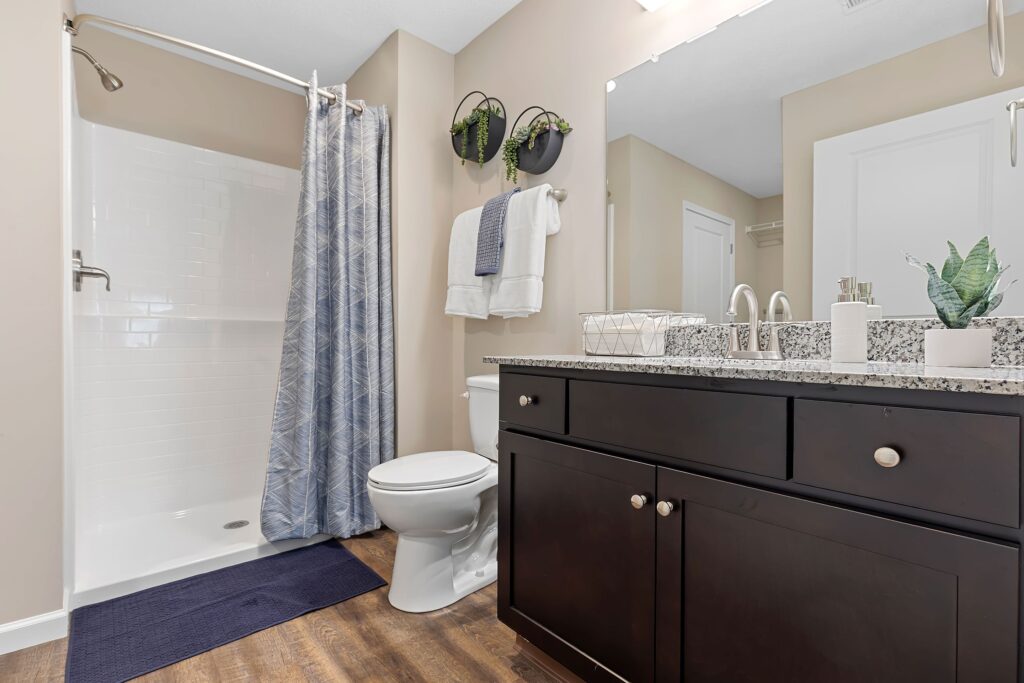 Modern bathroom with a walk-in shower, toilet, dark wood vanity with granite countertop, towels, wall-mounted plants, and a blue bath mat on wood-look flooring.