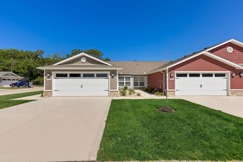 Single-story duplex with beige and red siding, two white double garages, concrete driveways, and well-kept lawns under a clear blue sky.