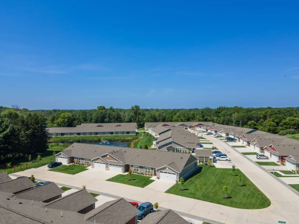 Aerial view of a residential neighborhood with single-story houses, green lawns, driveways, and parked cars under a clear blue sky, surrounded by trees.
