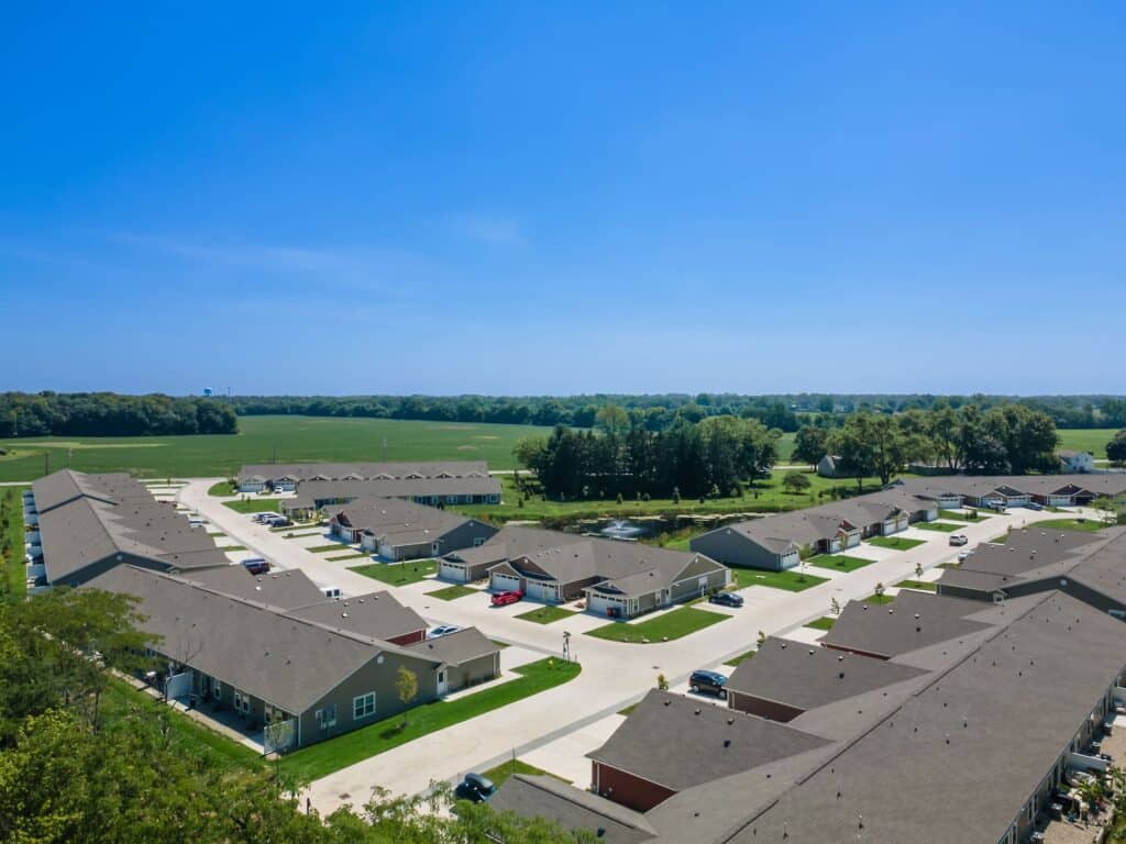 Aerial view of a suburban residential neighborhood with single-story houses, driveways, parked cars, and open green fields in the background under a clear blue sky.