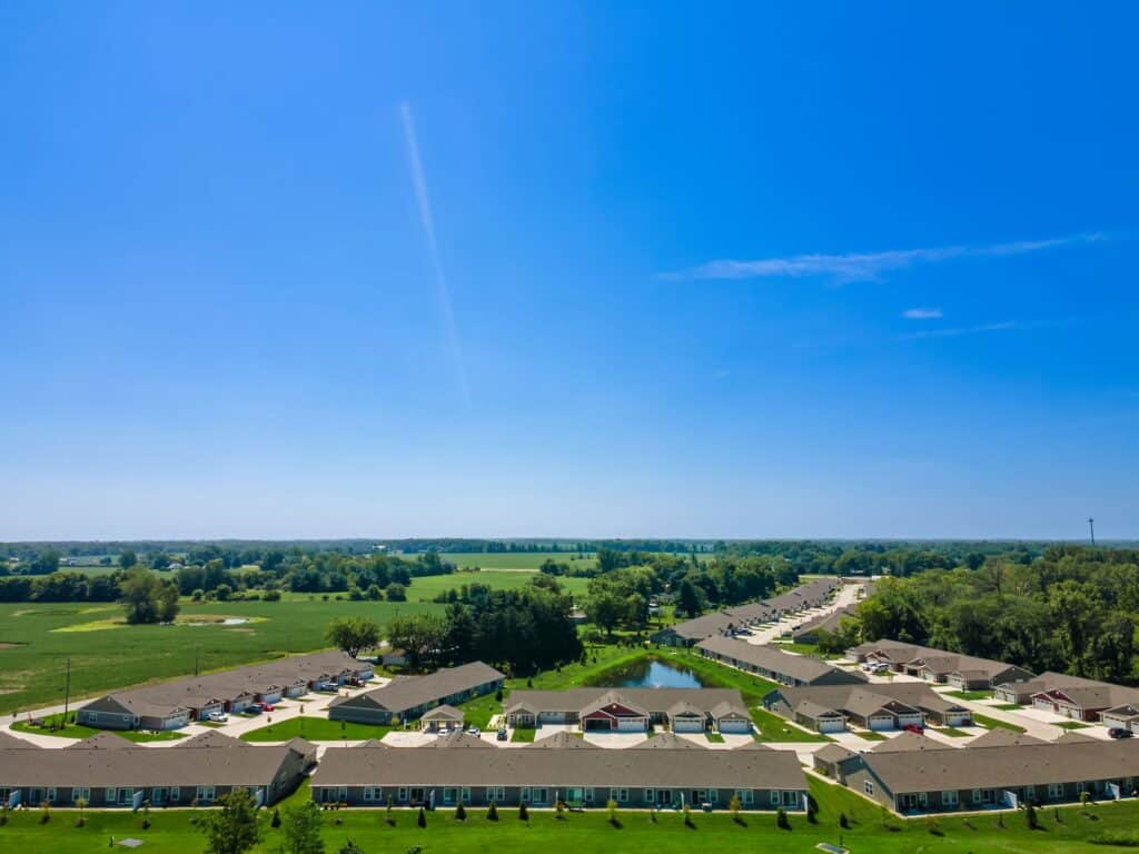 Aerial view of a residential neighborhood with single-story houses, a small pond, green lawns, and surrounding trees under a clear blue sky.