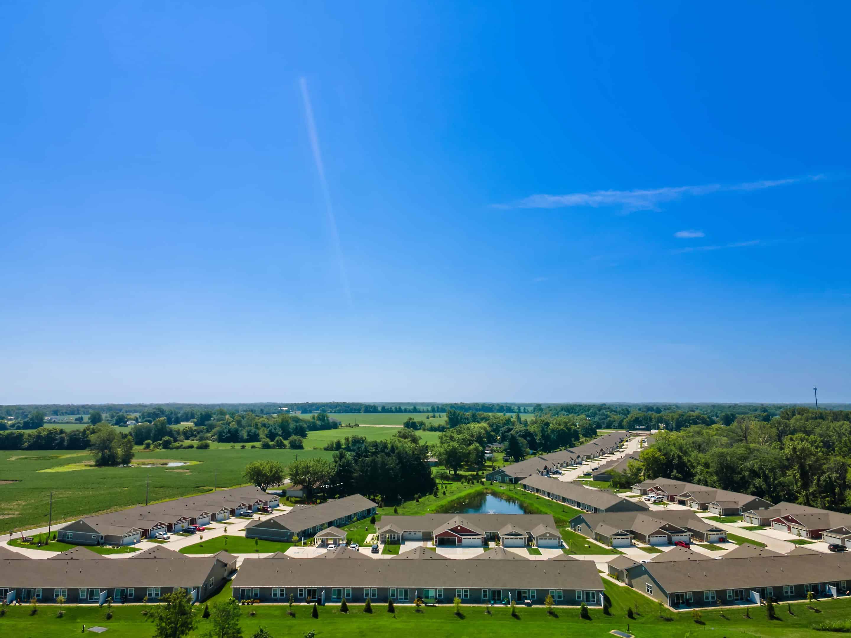 Aerial view of a residential neighborhood with single-story houses, a small pond, green lawns, and surrounding trees under a clear blue sky.
