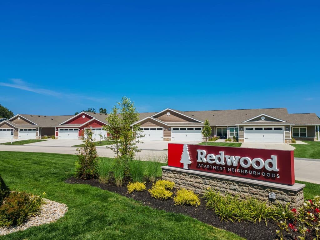 Redwood Apartment Neighborhoods sign in front of a row of single-story houses with garages, landscaped lawns, and a clear blue sky.