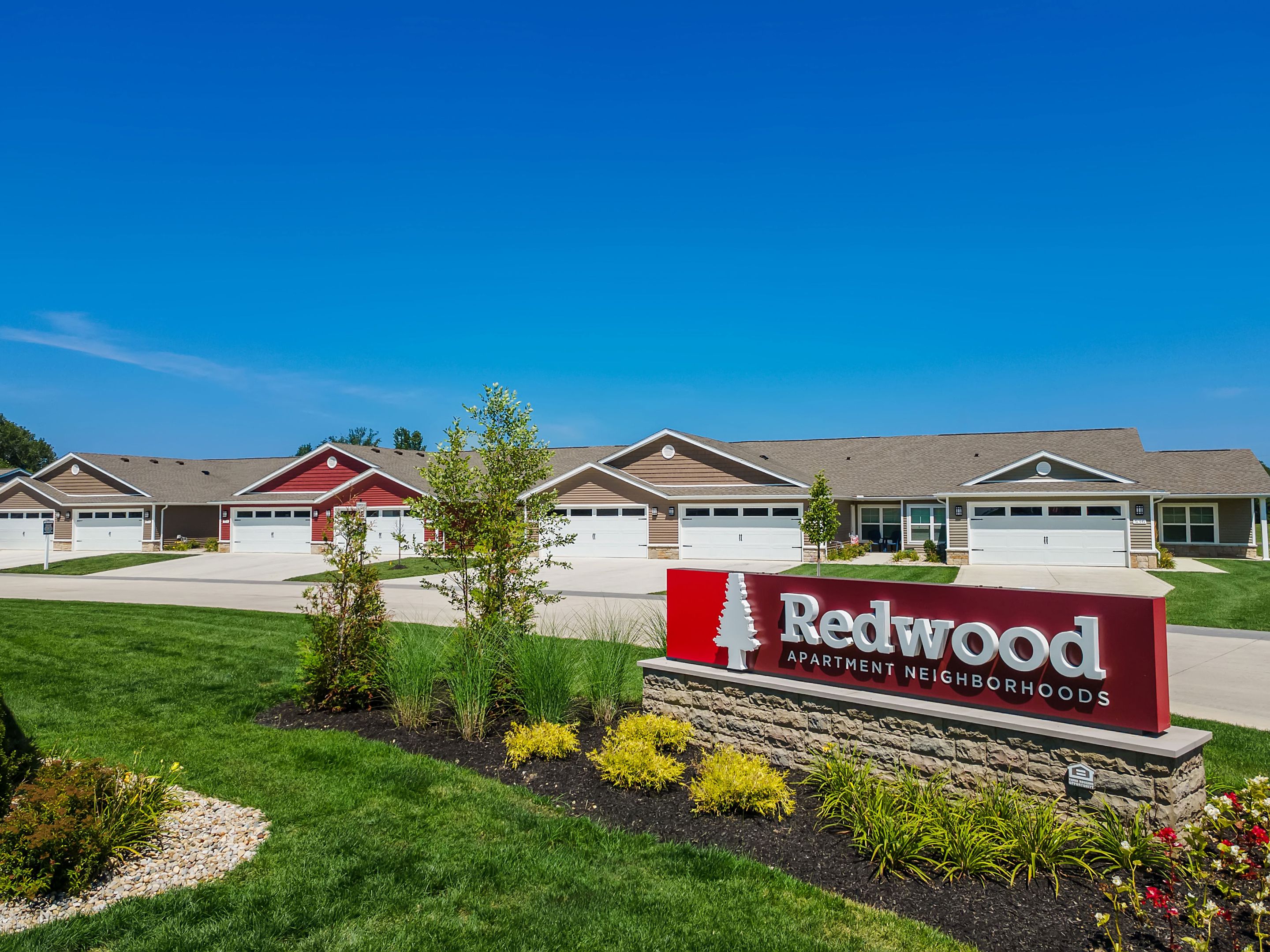 Redwood Apartment Neighborhoods sign in front of a row of single-story houses with garages, landscaped lawns, and a clear blue sky.