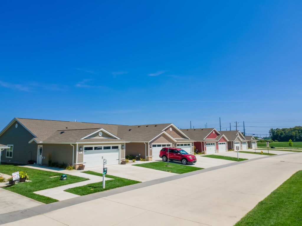Row of suburban single-story houses with garages, well-kept lawns, and a red car parked in one driveway under a clear blue sky.