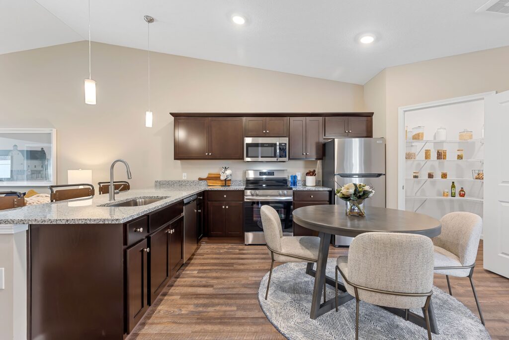 Modern kitchen with dark wood cabinets, stainless steel appliances, round dining table with four chairs, and open pantry stocked with baskets and jars.