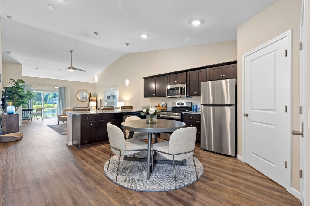 Open-concept kitchen and dining area with dark wood cabinets, stainless steel appliances, round table with four chairs, and wood flooring. Living area and patio visible in the background.