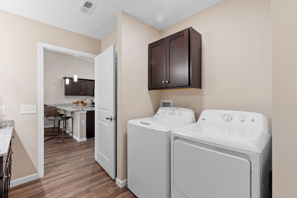 Laundry room with a white washing machine and dryer, brown wall cabinets above, and a view into a kitchen with bar stools and pendant lights.