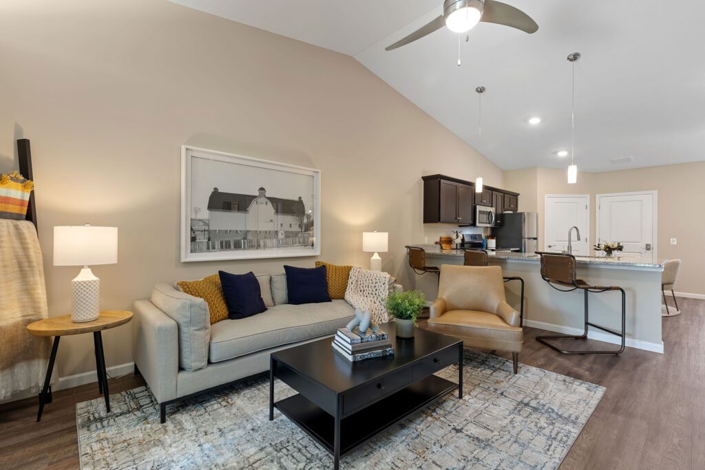 Modern living room and kitchen with beige walls, a sofa, an armchair, a coffee table, and a counter with bar stools under pendant lights.