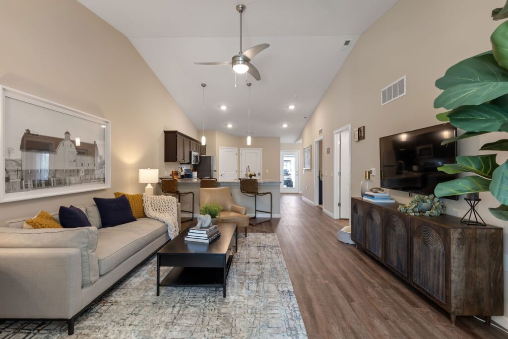 Modern open-concept living room and kitchen with beige walls, wood floors, a sofa, coffee table, TV console, and kitchen island with stools under ceiling fans and recessed lighting.