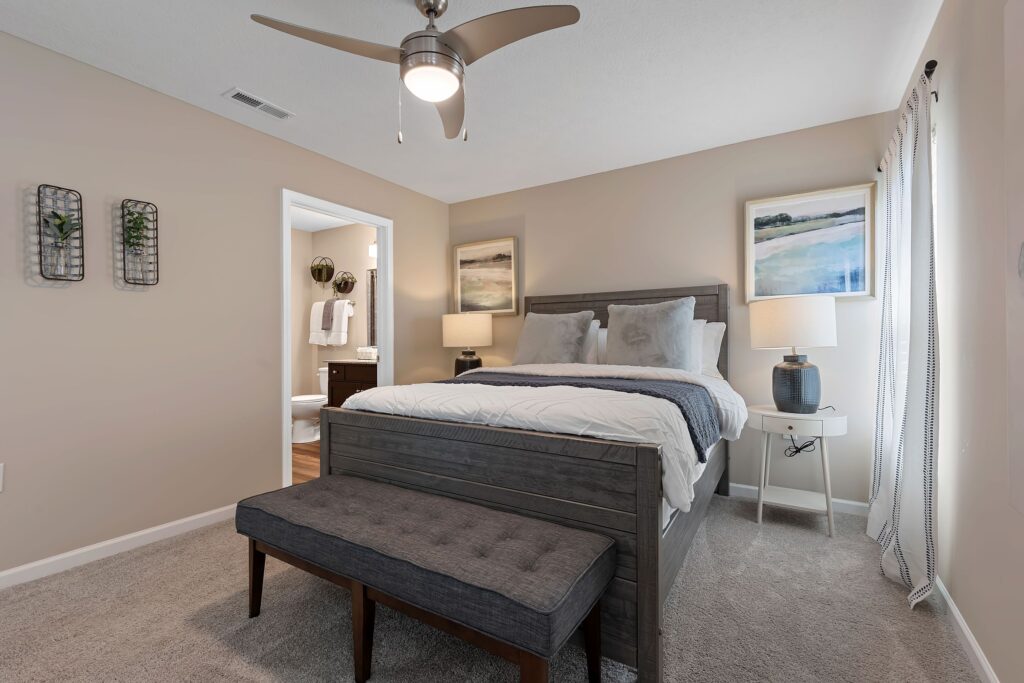 Modern bedroom with a gray bed, bench, two bedside tables with lamps, neutral walls, carpeted floor, and an open door leading to a bathroom.