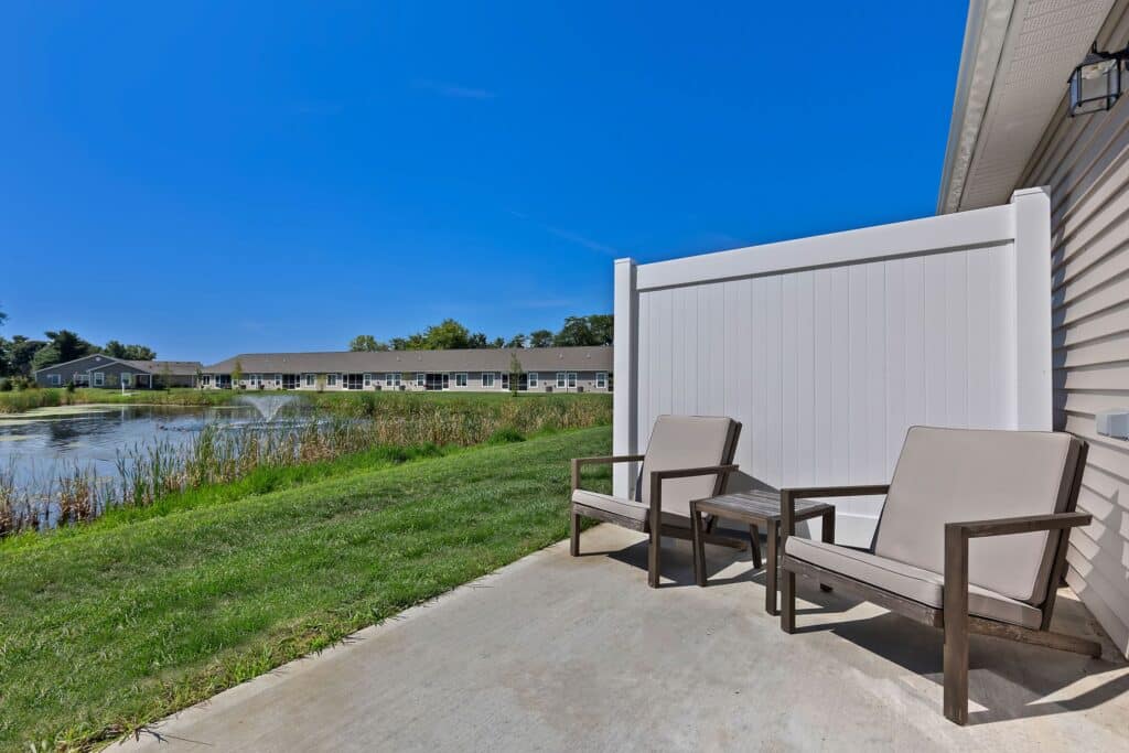 Two patio chairs and a small table on a concrete patio overlook a pond and grassy area, with single-story buildings in the background under a clear blue sky.