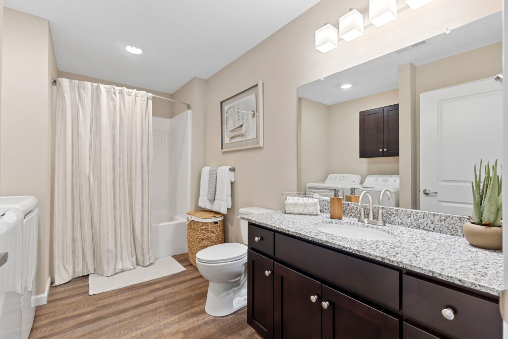 Modern bathroom with granite countertop, dark wood cabinets, a white toilet, shower with curtain, washer and dryer, and neutral-colored walls and decor.