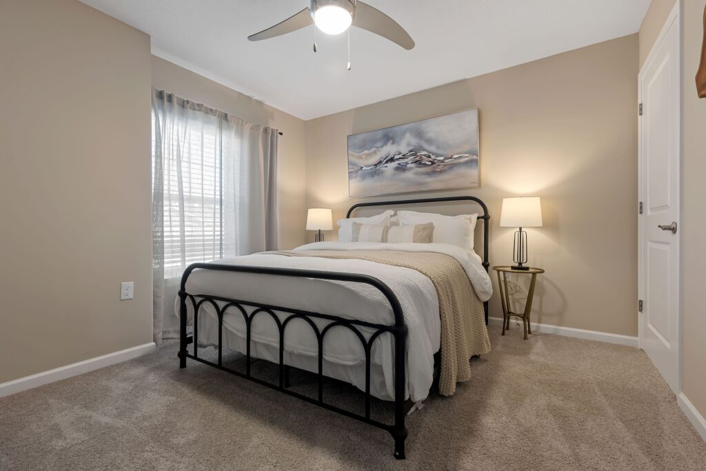 A neatly made bed with a metal frame sits between two nightstands with lamps in a beige-carpeted bedroom with tan walls and a window with sheer curtains.