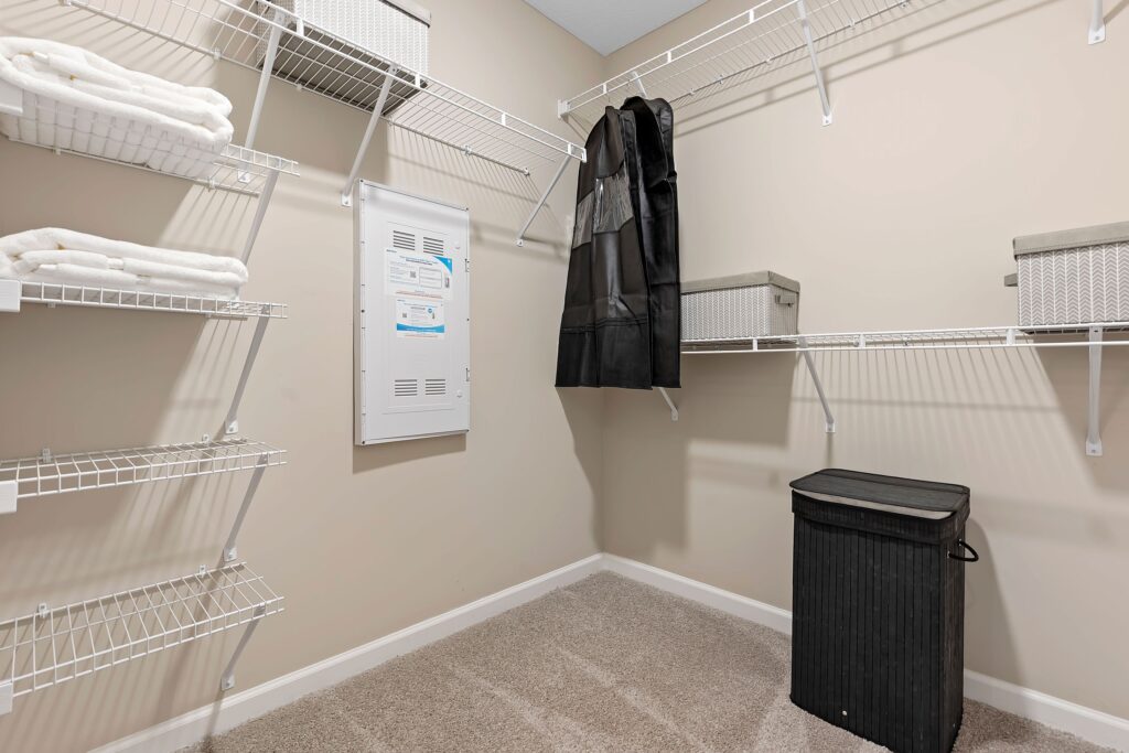 A walk-in closet with wire shelving, folded towels, storage bins, a hanging garment bag, a laundry hamper, and an electrical panel on a beige wall.