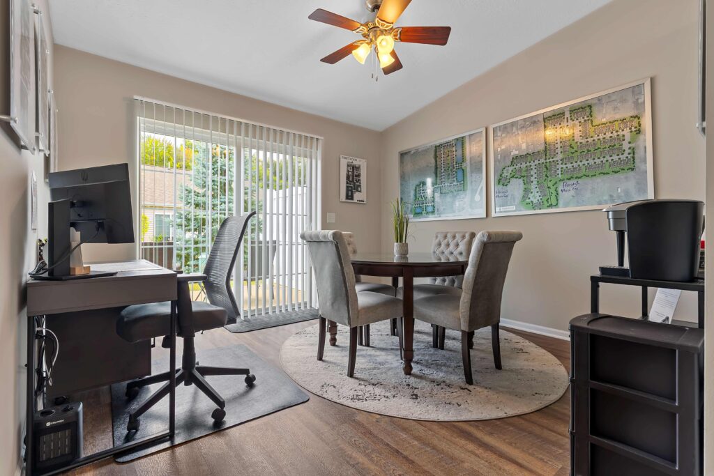 A home office with a desk, computer, four upholstered chairs around a round table, wall art, a ceiling fan, and sliding glass doors letting in natural light.