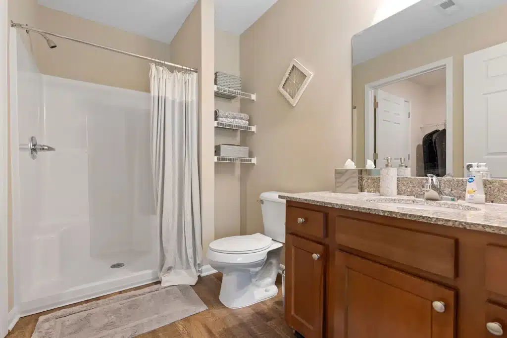 Bathroom with a walk-in shower, curtain, toilet, shelves with baskets, wooden vanity with granite countertop, and a wall mirror.