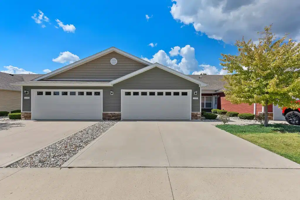 A duplex with two double garages, tan siding, and stone accents, separated by a rock and concrete divider; a small tree and green grass are visible on the right side.