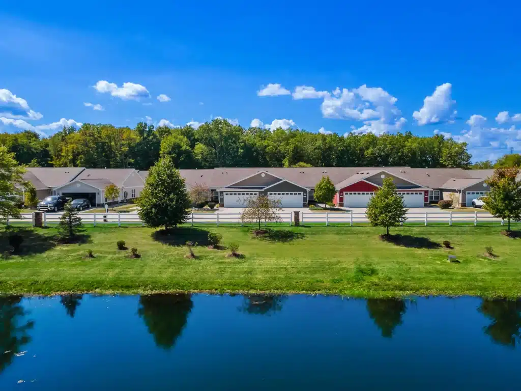 Row of single-story suburban houses with garages, separated by a white fence, facing a grassy area and pond under a blue sky with scattered clouds.