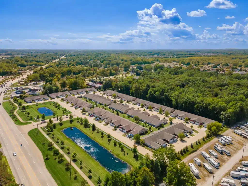 Aerial view of a suburban residential complex with rows of houses, two retention ponds, a forested area, and RVs parked along one side, bordered by a main road.