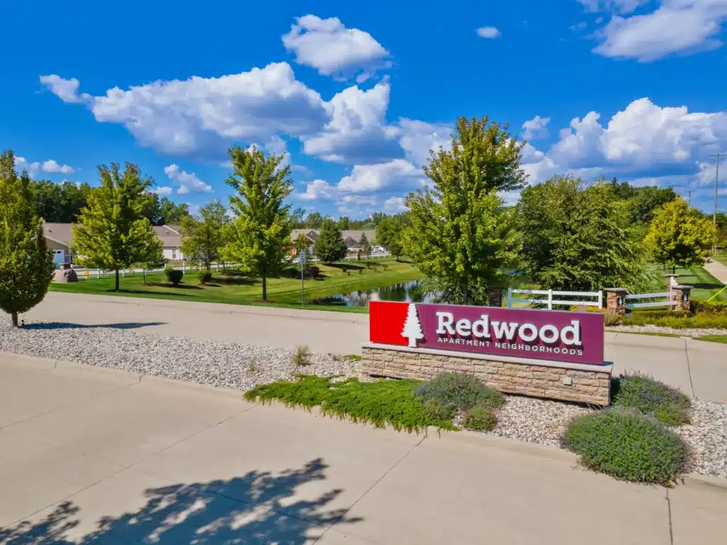 Redwood Apartment Neighborhoods sign on a landscaped median with trees, houses, and a pond visible in the background under a blue sky with scattered clouds.