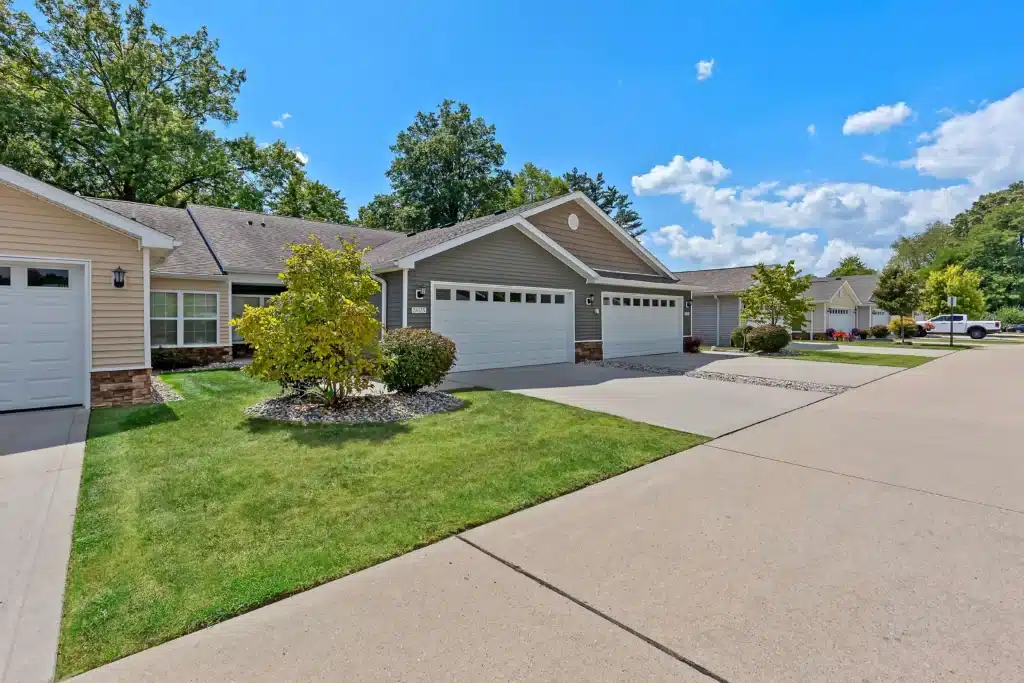 Single-story townhomes with attached garages and well-maintained lawns on a sunny day. Concrete driveway and trimmed landscaping are visible.