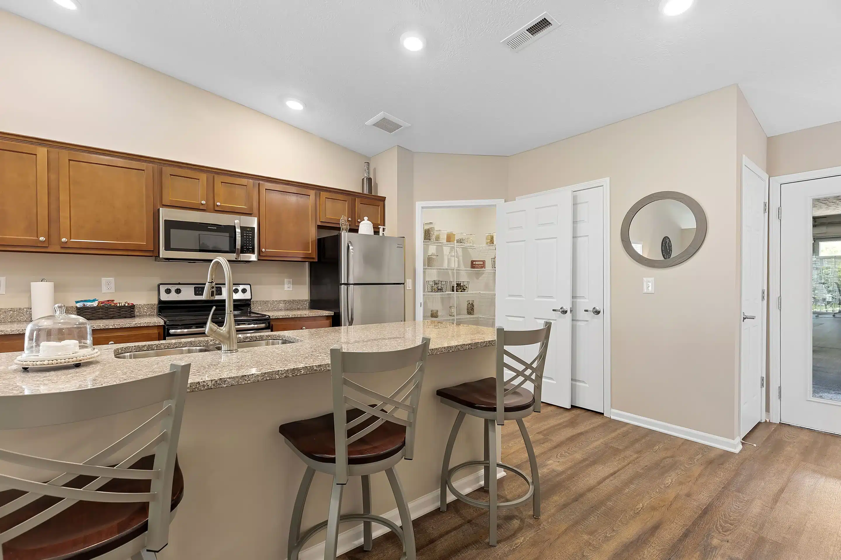 Modern kitchen with brown cabinets, stainless steel appliances, granite countertops, and three bar stools at a breakfast bar. Pantry and glass door visible in the background.