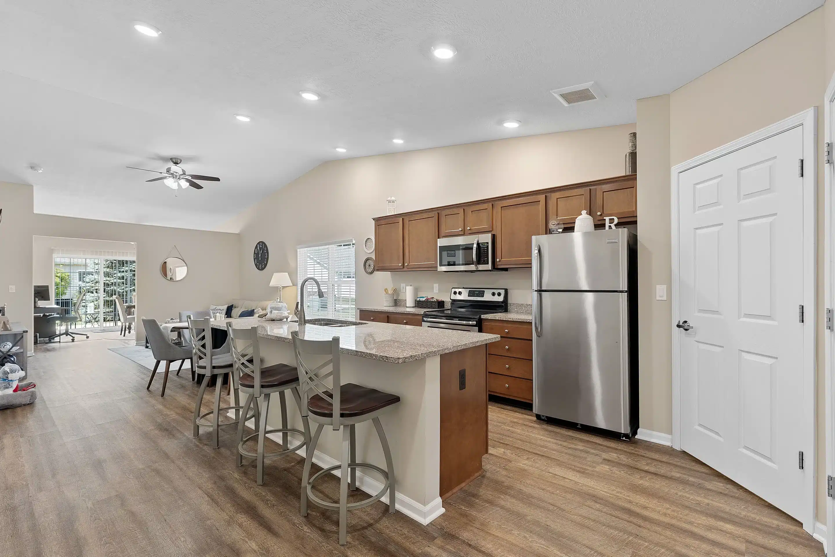 Modern kitchen with wooden cabinets, stainless steel appliances, an island with bar stools, and an open layout connecting to a dining and living area with wood flooring.